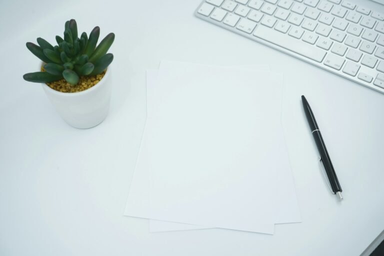 Blank paper, a pen, a white keyboard and a small plant on a light desk