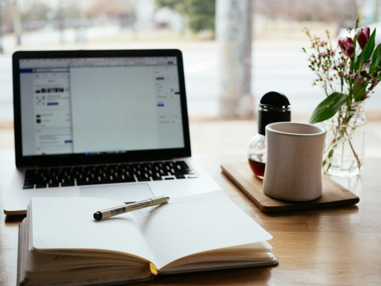 Laptop beside an open notebook, pen and mug near a window